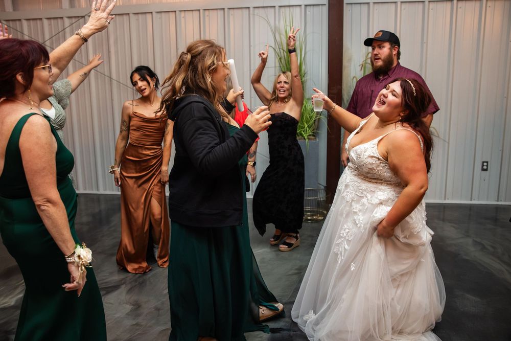 Wedding guests in formal dresses dance with raised arms at a reception, with a bride in a white gown smiling in the foreground.
