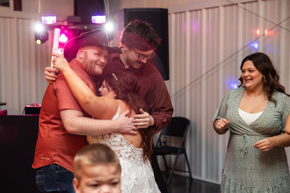 Guests embrace in a warm group hug on a dance floor at a festive wedding reception, with a smiling woman and a child nearby.