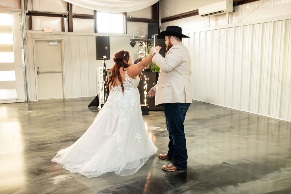 Bride in a flowing white gown and groom in a cowboy hat share a first dance on a polished concrete floor at a rustic wedding venue.