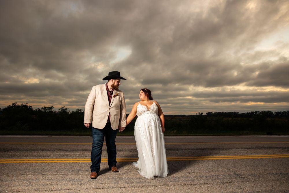 Bride in a white gown and groom in a cream blazer and black cowboy hat hold hands on a empty road under a dramatic cloudy sunset sky.