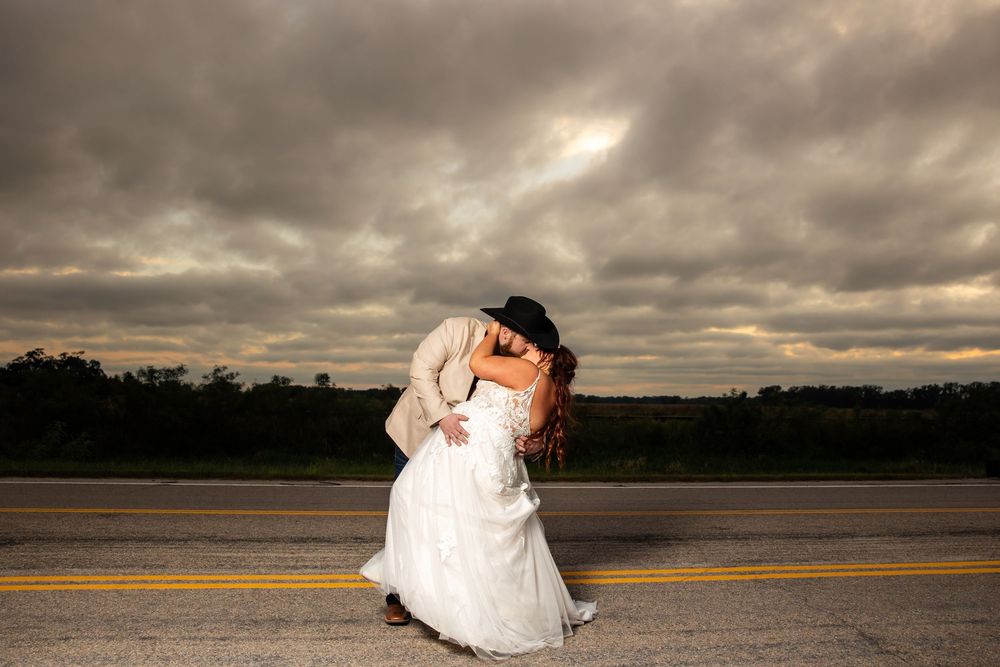 Bride in a white gown and groom in a cowboy hat share a dramatic dip kiss on a country road beneath a moody, cloudy sky.