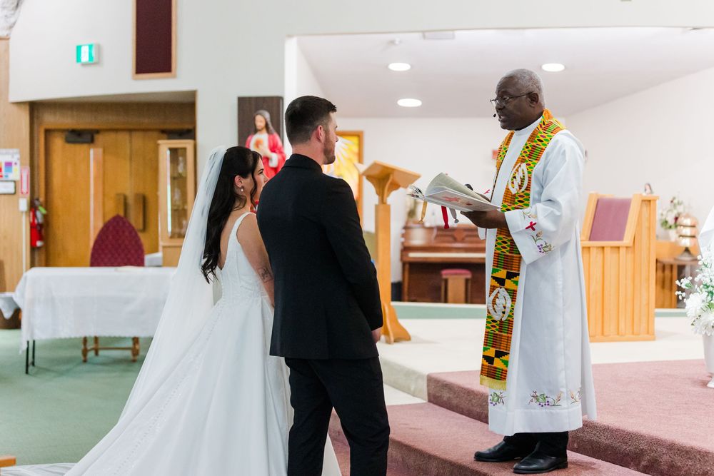 Bride in a white gown and groom in a dark suit stand before a priest in vestments holding a book during a wedding ceremony in a church.