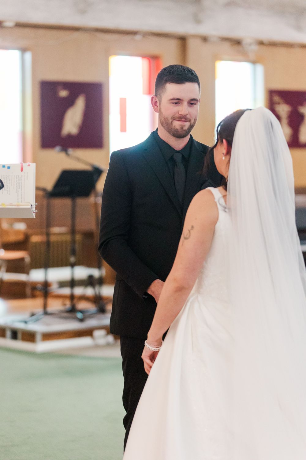 A smiling groom in a black suit and tie faces his bride in a white gown and long veil during a wedding ceremony inside a church.