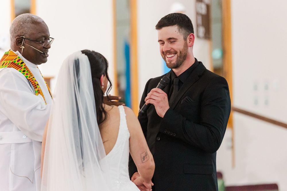Smiling groom in a black suit holds a microphone while exchanging wedding vows with a bride in a veil as an officiant looks on.