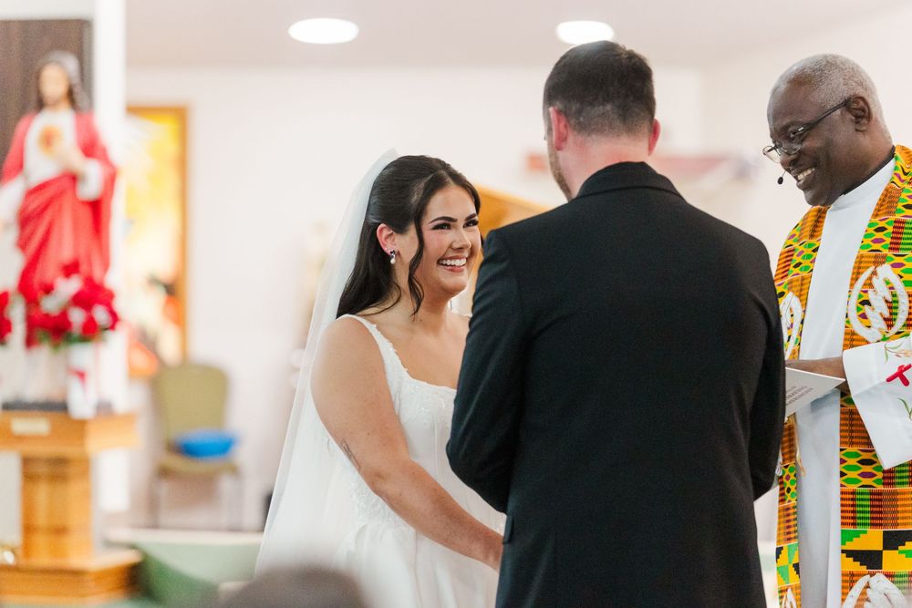 Smiling bride in a white gown holds hands with the groom during a church wedding ceremony led by an officiant in a colorful kente stole.