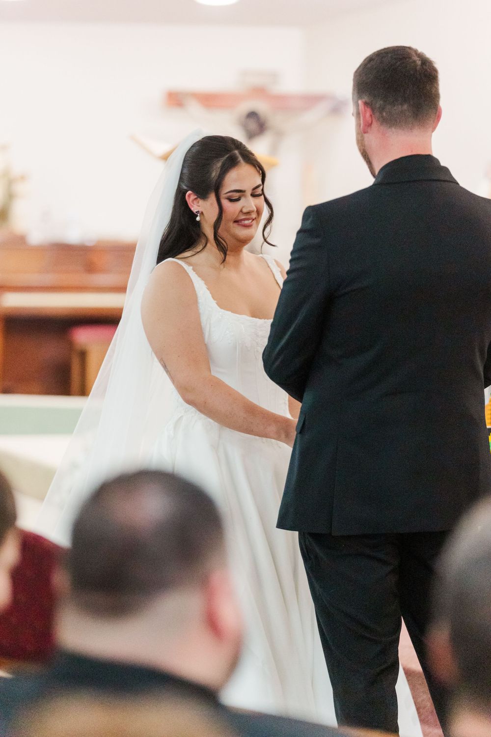 Smiling bride in a white gown and veil faces the groom in a dark suit during a wedding ceremony in a church as guests look on.