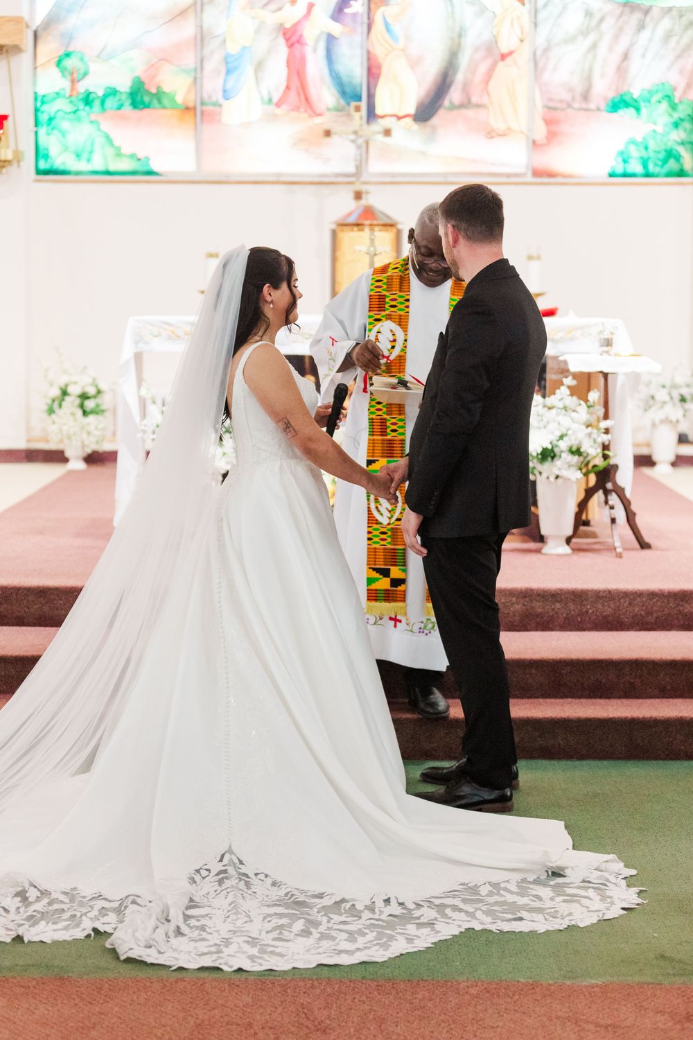 Bride in a white gown with a long lace-trimmed veil and groom in a black suit exchange vows before an officiant at a church altar.