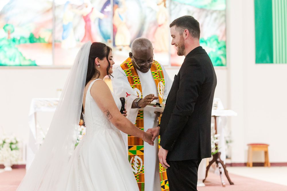 Bride in a white gown and groom in a black suit hold hands before an officiant in colorful kente stole during a church wedding ceremony.