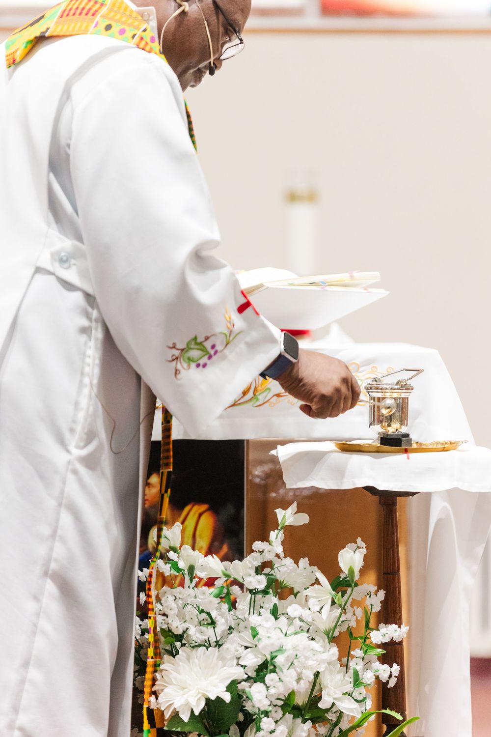 Clergy member in white embroidered vestments with a colorful stole prepares the altar beside white flowers during a church service.