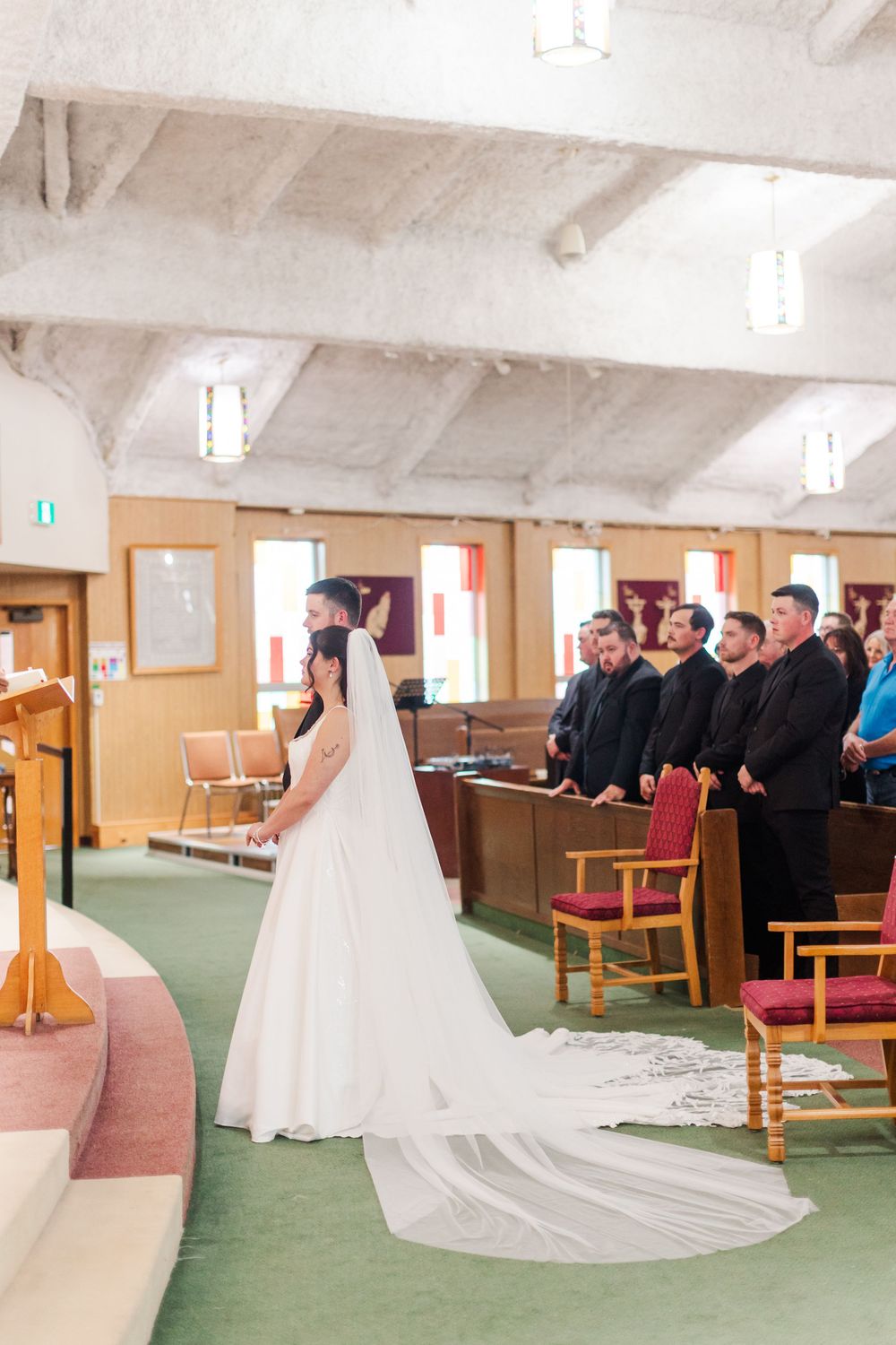 Bride in a white gown with a long veil and lace train stands at the altar during a church wedding as guests in dark suits look on.