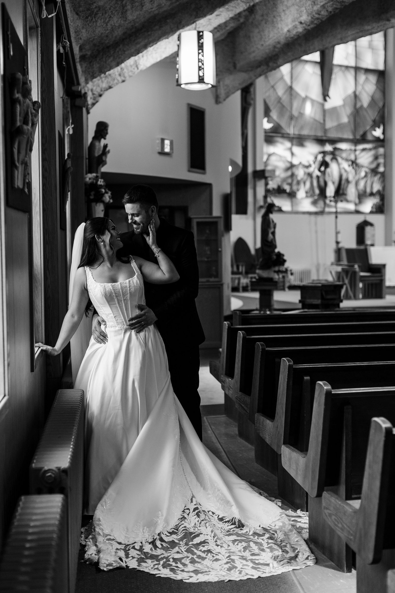 Black and white photo of a bride in a lace-trimmed gown embracing her groom beside church pews with stained glass windows behind them.