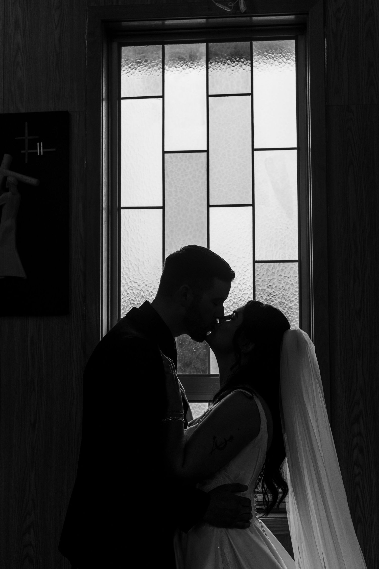 Black and white silhouette of a bride and groom sharing a kiss in front of a stained glass window during their wedding.