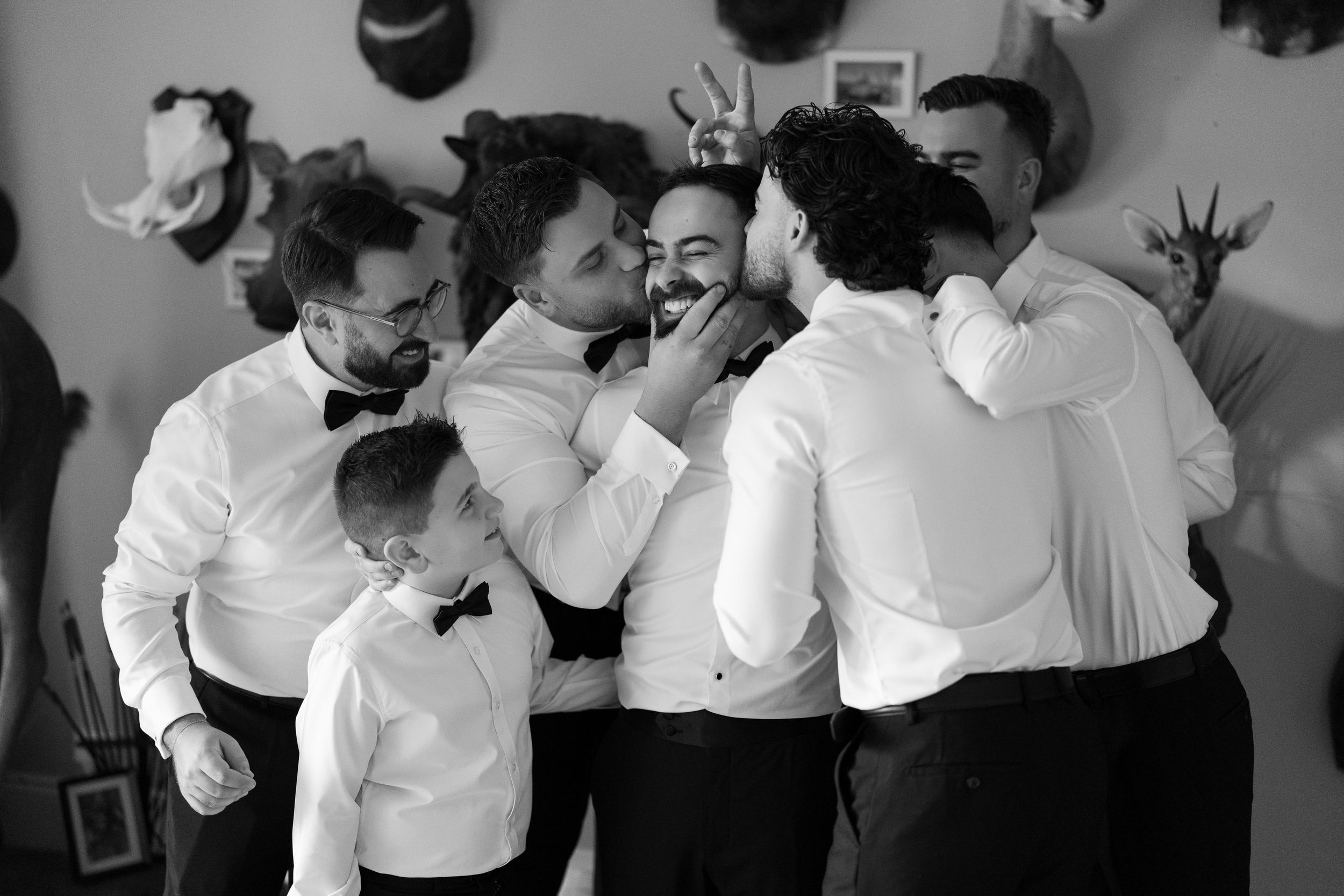 Black and white photo of groomsmen in bow ties and white shirts joyfully hugging and kissing the groom while a young boy smiles beside them.