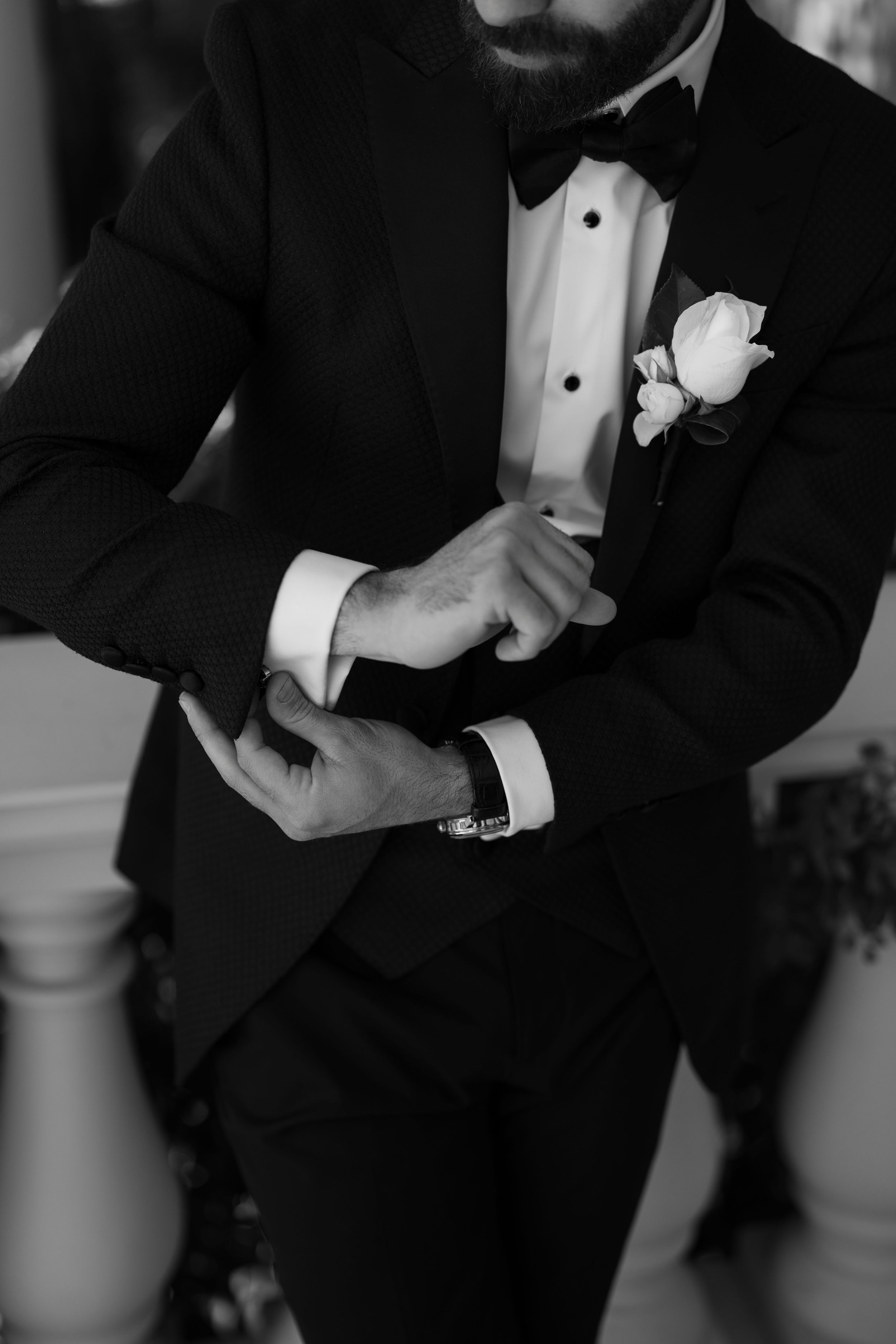 Black and white photo of a groom in a tuxedo and bow tie with a white rose boutonniere adjusting his shirt cuff and wristwatch.