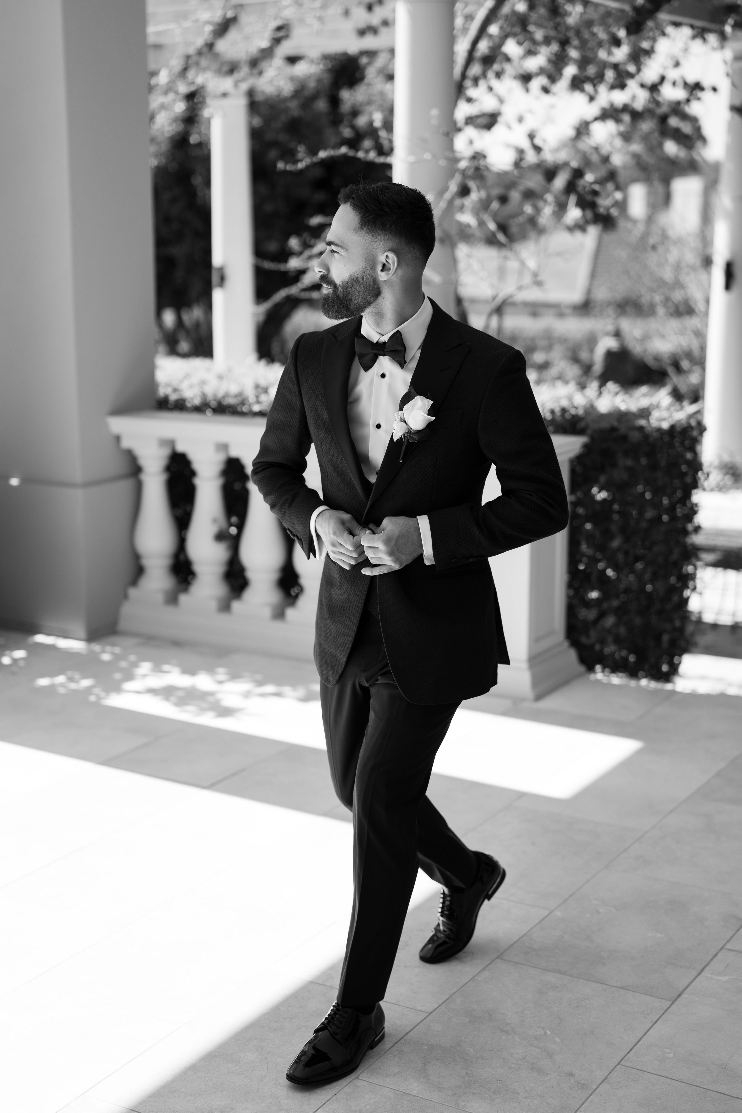 Groom in a black tuxedo, bow tie and white boutonniere adjusts his jacket while walking across a sunlit stone terrace in black and white.
