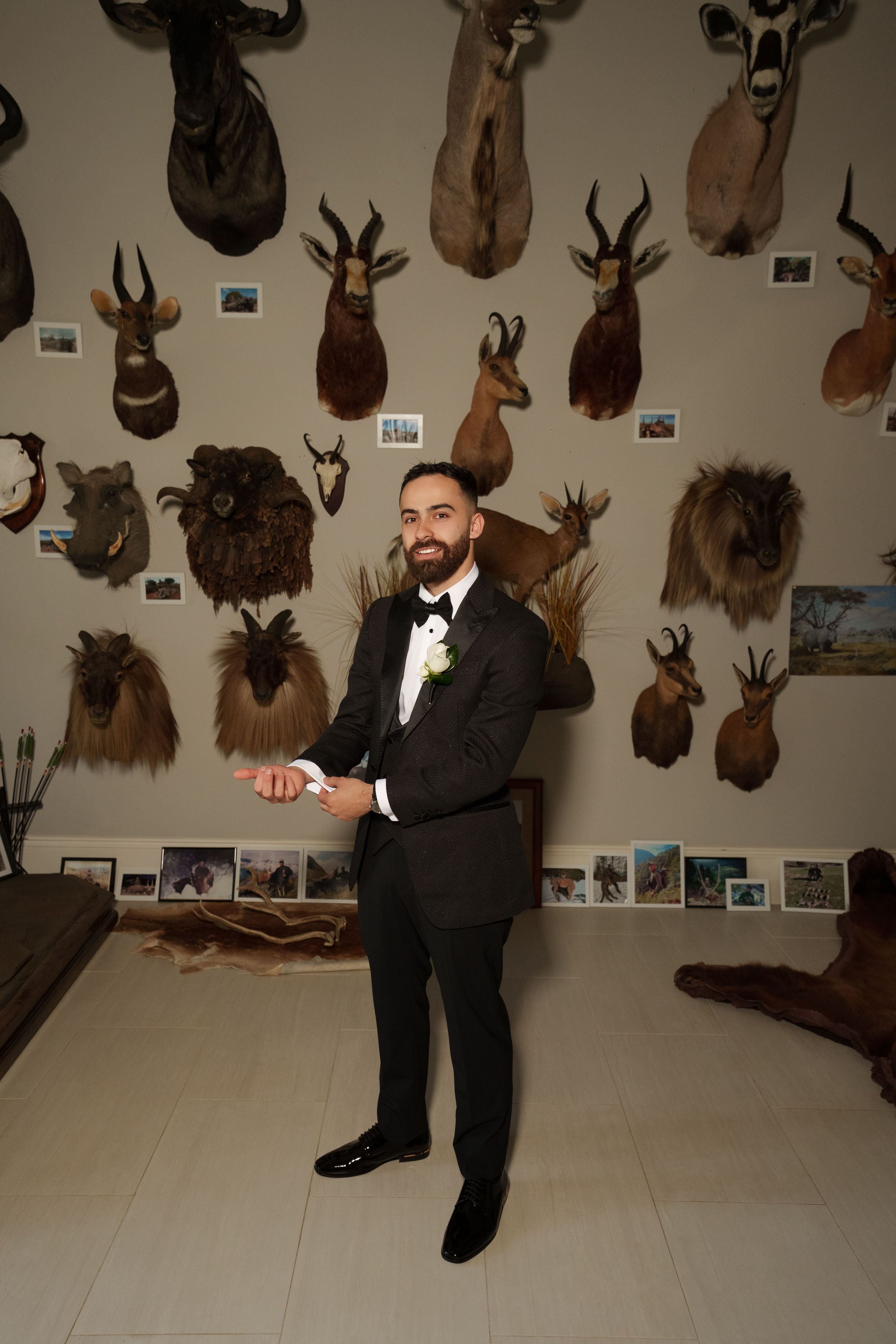 Groom in black tuxedo and bow tie adjusts his cuff before a wall of mounted animal trophy heads and framed photos.