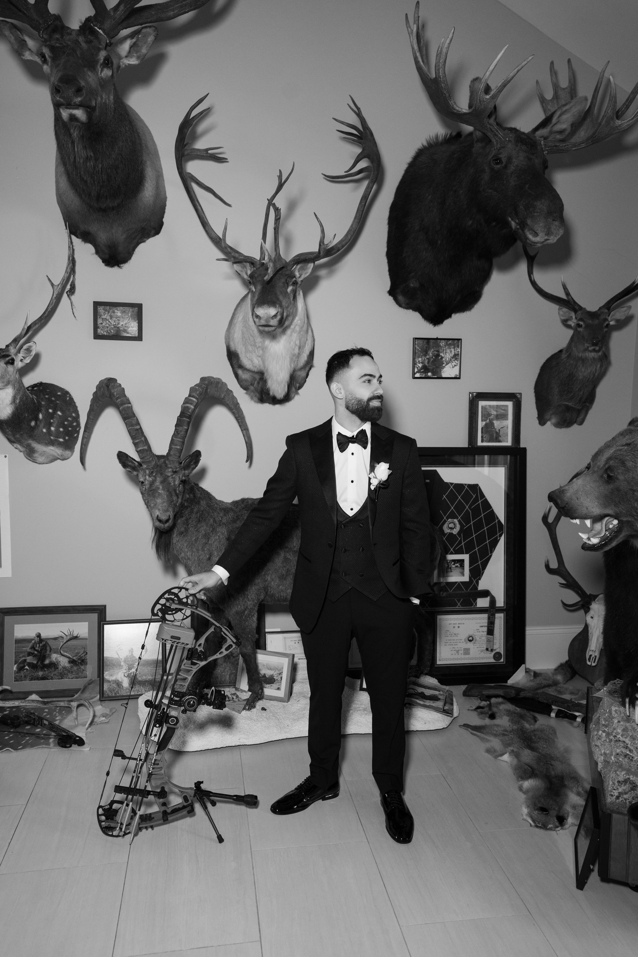 Groom in a black tuxedo and bow tie holds a compound bow in a room decorated with mounted deer, elk, moose and ibex trophy heads.