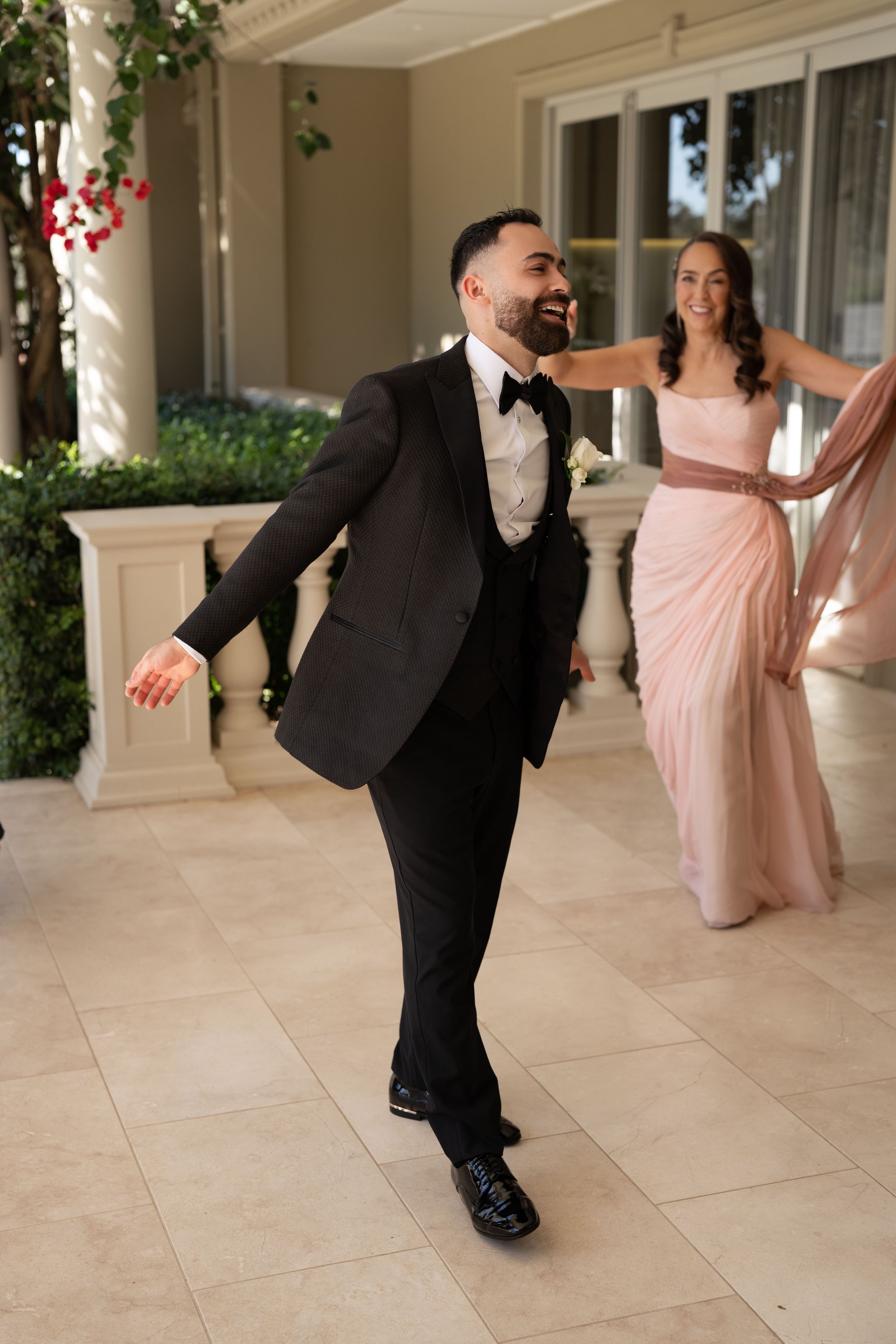 Smiling groomsman in a black tuxedo and bow tie walks joyfully on a stone terrace while a woman in a blush pink gown dances behind him.