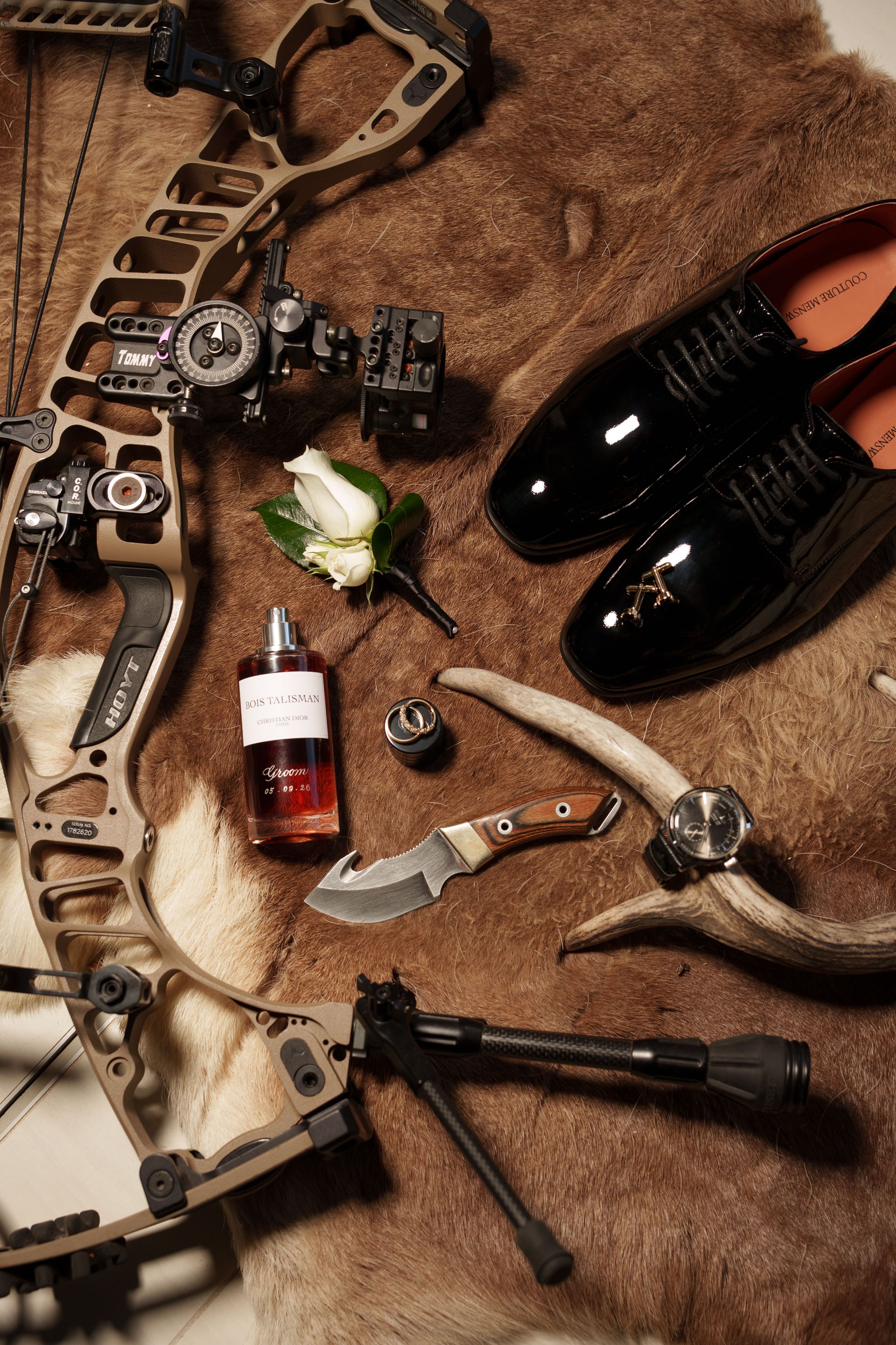 Flat lay of a compound bow, patent leather dress shoes, cologne bottle, hunting knife, white boutonniere and antler on a fur hide.