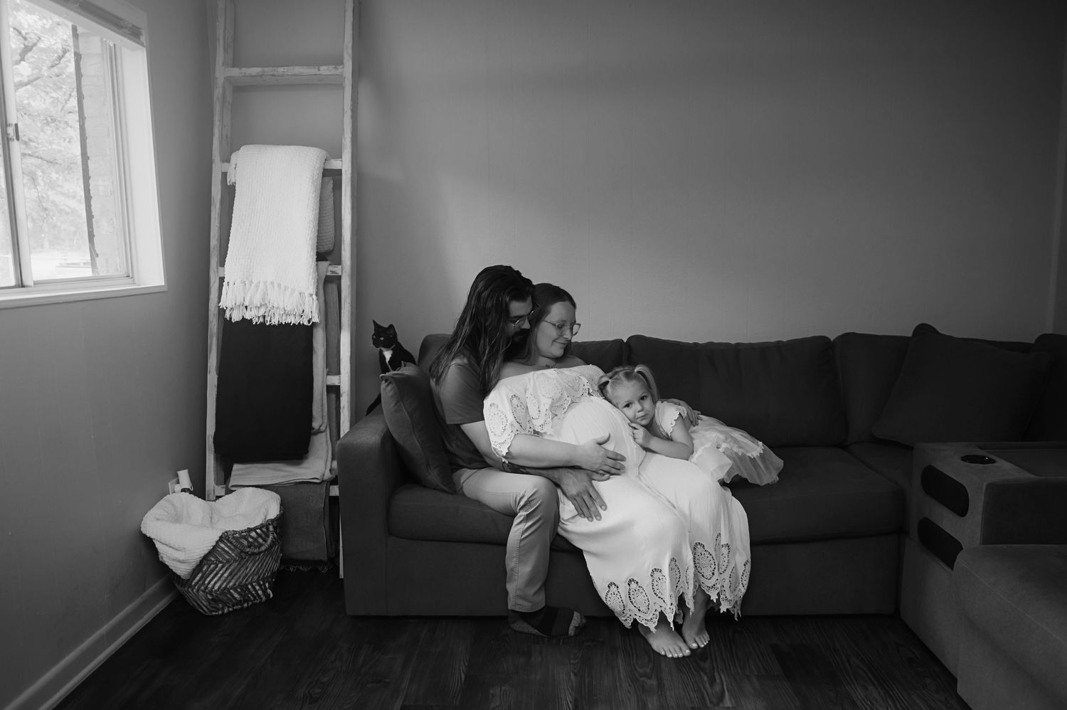 Black and white photo of a mother cradling her baby in a white lace gown on a cozy living room sofa beside a window.