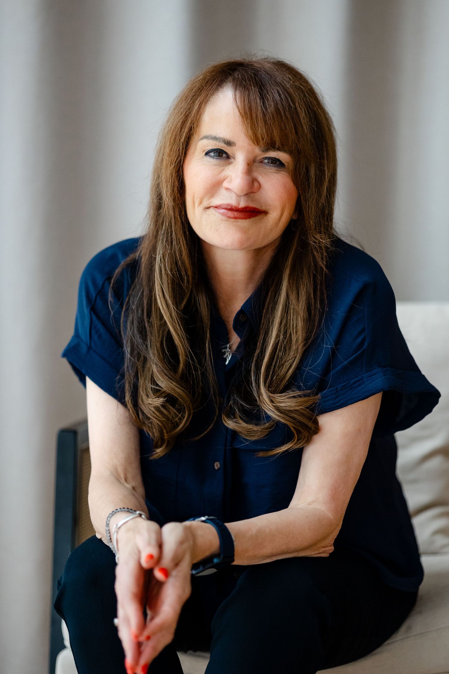A smiling woman with long brown hair wearing a navy blue top sits relaxed on a white chair in a bright room.