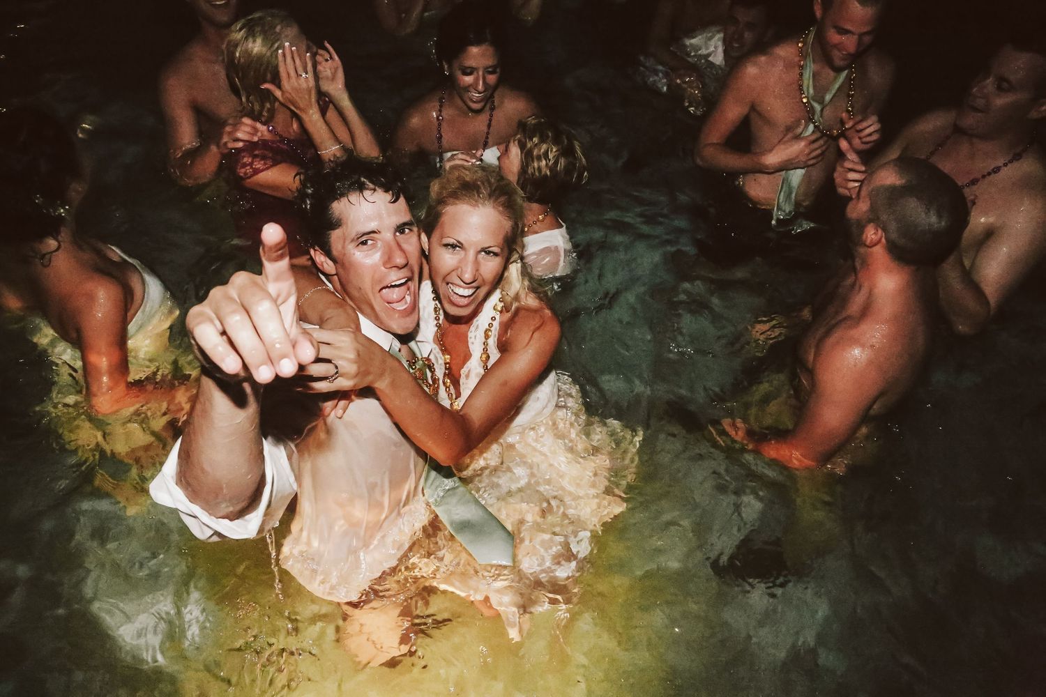 Couple celebrating in a pool during their wedding reception, surrounded by guests in formal attire splashing in water.