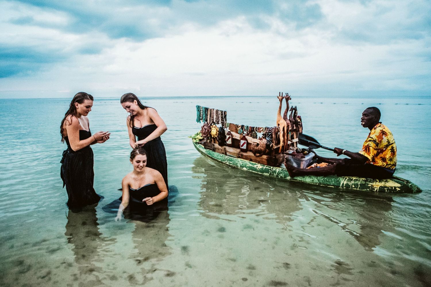 Three women in black dresses wade in shallow ocean water near a colorful boat rowed by a local man on a tropical beach.
