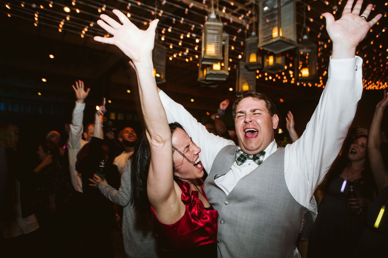 Joyful couple dancing with hands raised at a festive wedding reception with string lights and lanterns.
