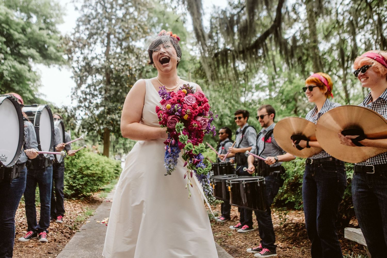 Bride in white gown laughing with colorful bouquet, surrounded by live band musicians outdoors at wedding celebration.