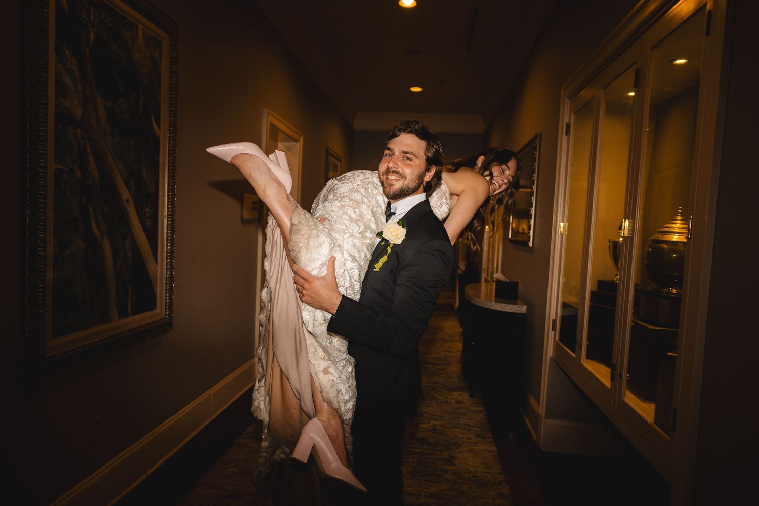 Groom in black tuxedo carrying bride in white wedding gown through a warmly lit hotel hallway.