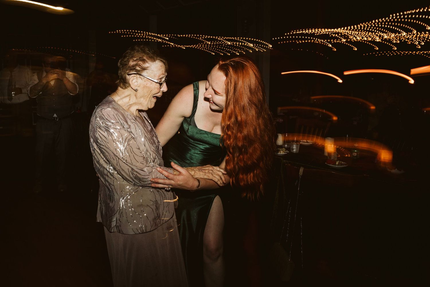 Two women share a joyful moment dancing together at a formal event, one elderly in lace and one in a green gown.
