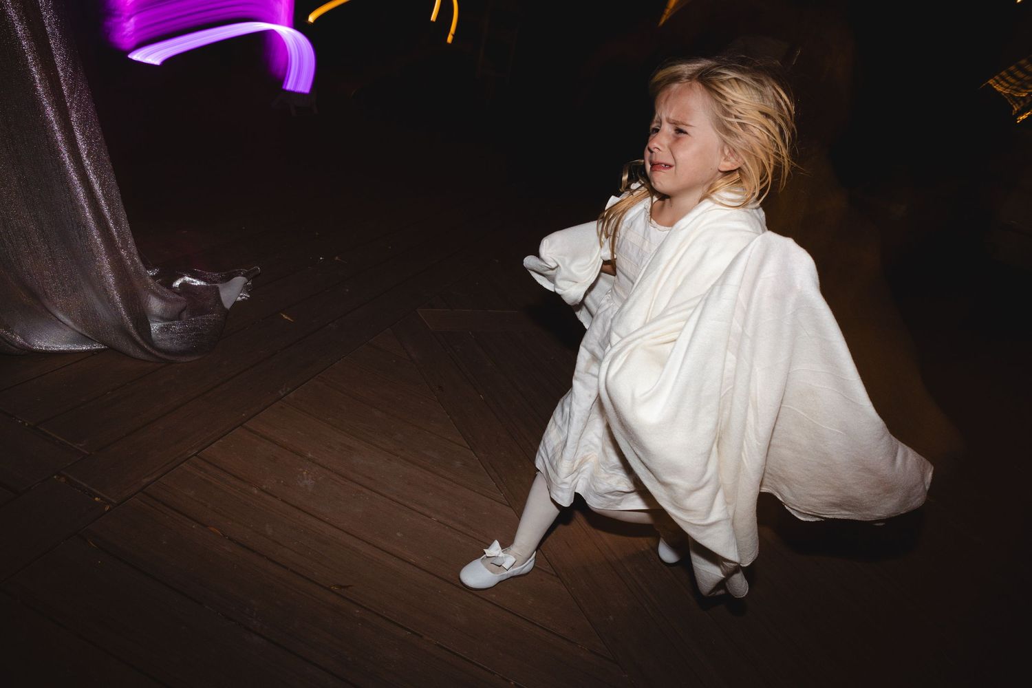 Young blonde girl in white dress sitting on wooden dance floor at a wedding reception with colorful lights.