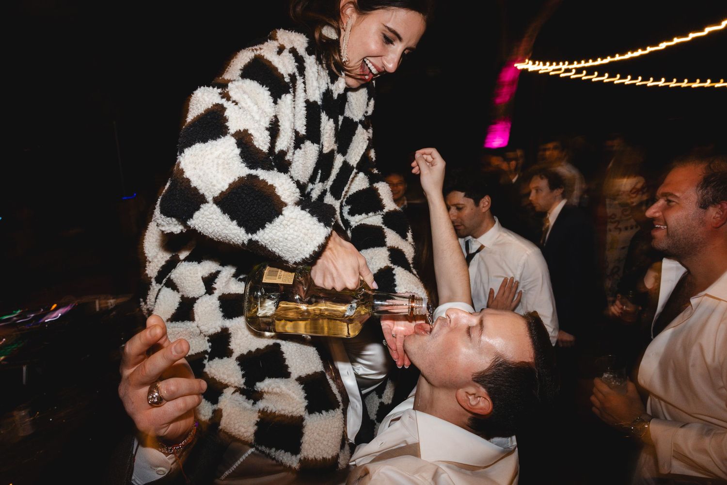 Woman in checkered sweater pours champagne into mans mouth on dance floor at lively party.