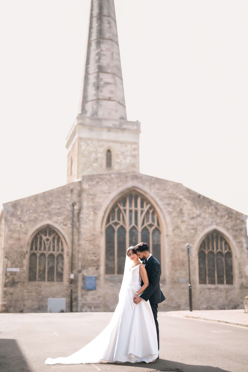 Bride and groom embrace in front of a historic stone church with gothic spire on their wedding day.