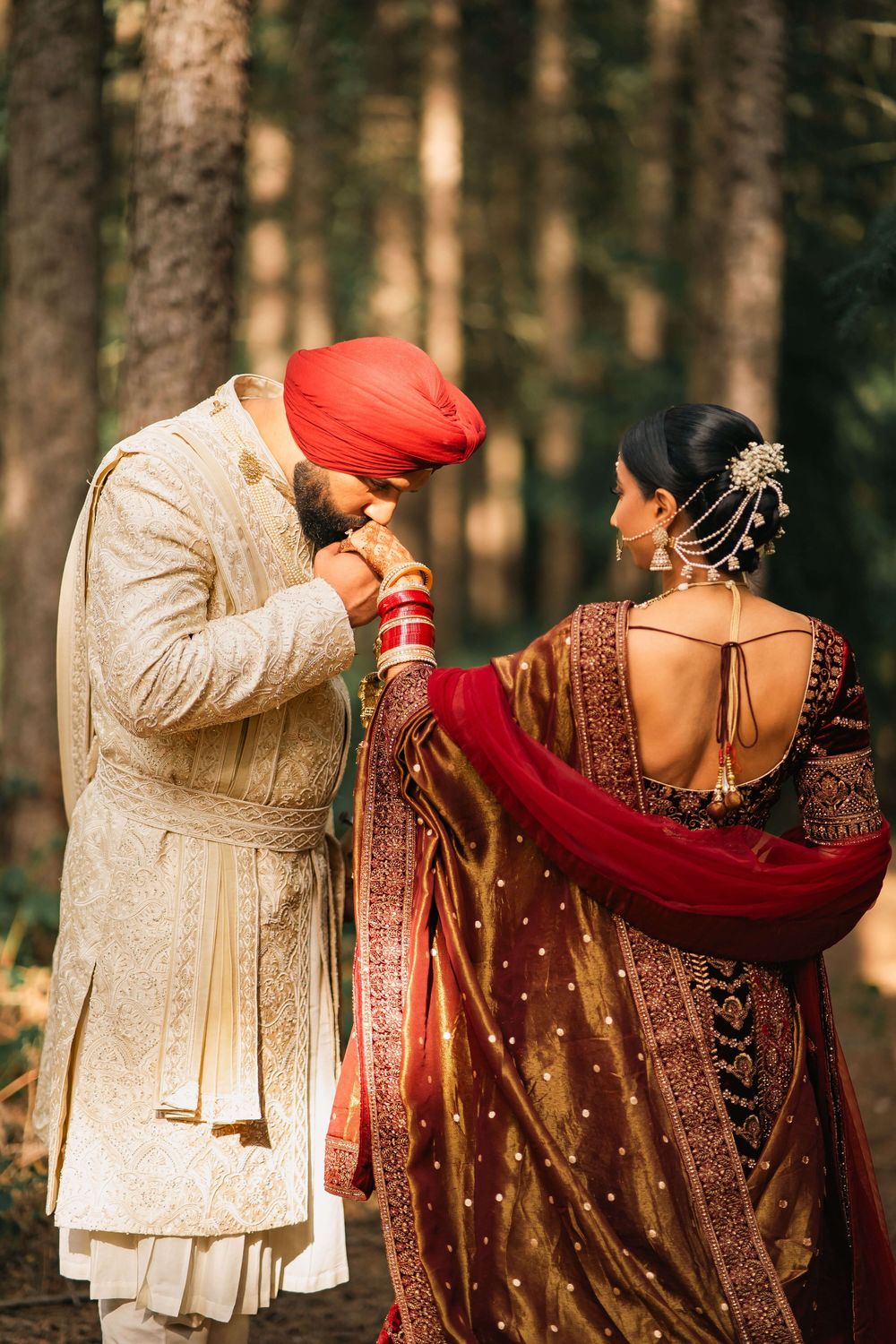 Sikh groom in red turban kissing bride's hand in forest, both wearing traditional Indian wedding attire.