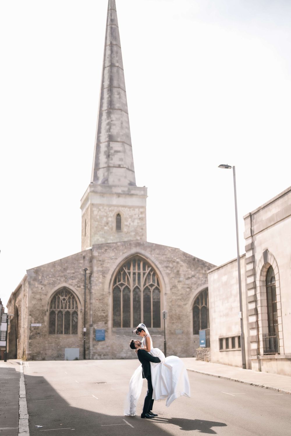 Groom lifting bride in wedding attire outside historic stone church with tall spire on sunny day.