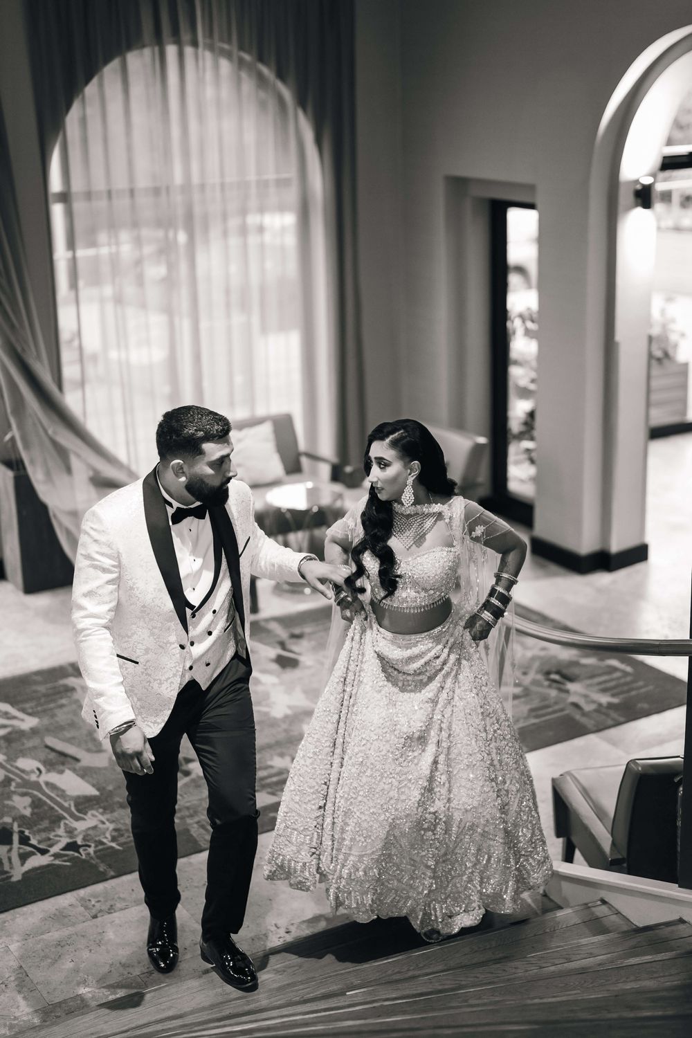 Black and white photo of a bride in embellished lehenga and groom in white tuxedo sharing a romantic moment.