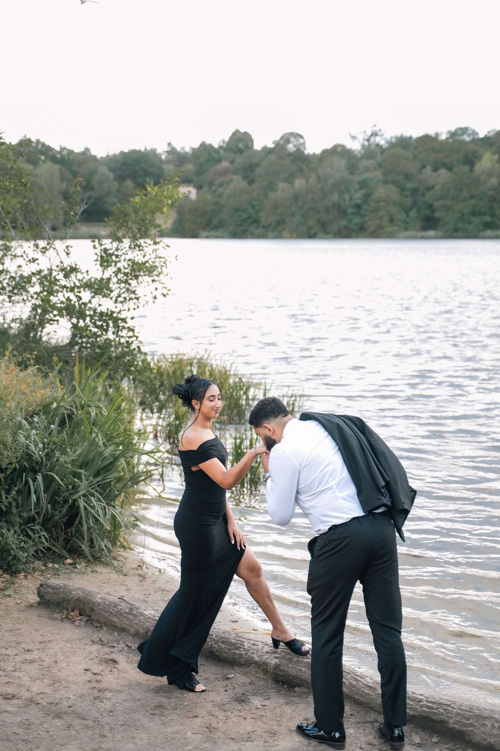 Elegant couple in formal attire sharing a romantic moment by a serene lake surrounded by lush green trees.