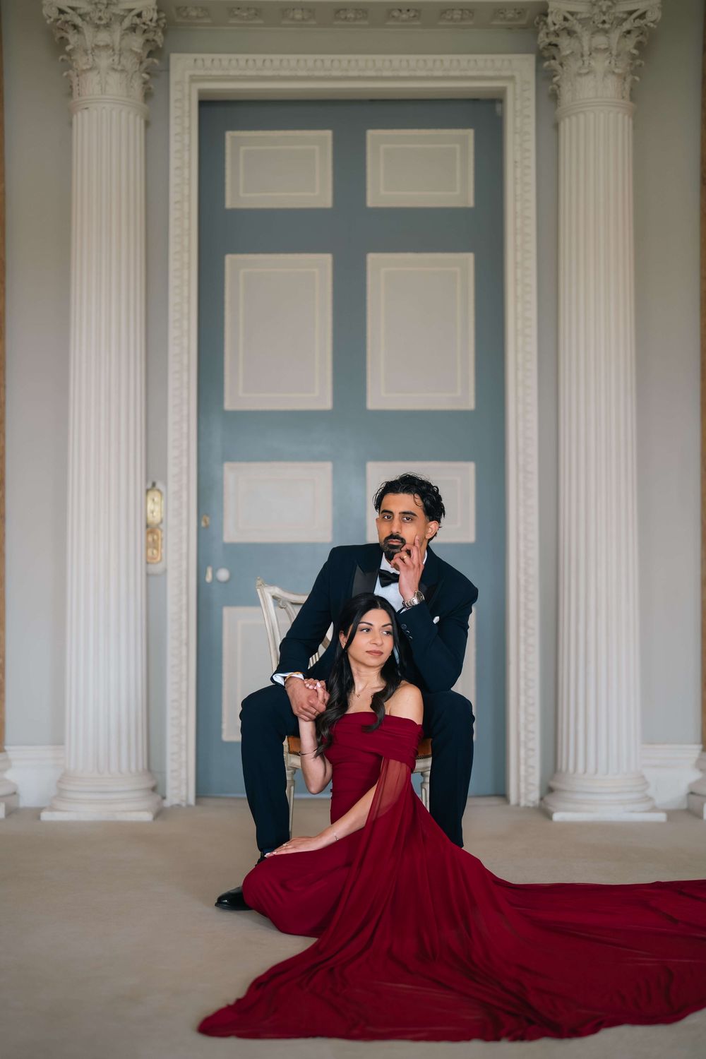 Couple posing elegantly in front of blue door, woman in red gown seated, man in tuxedo behind her.