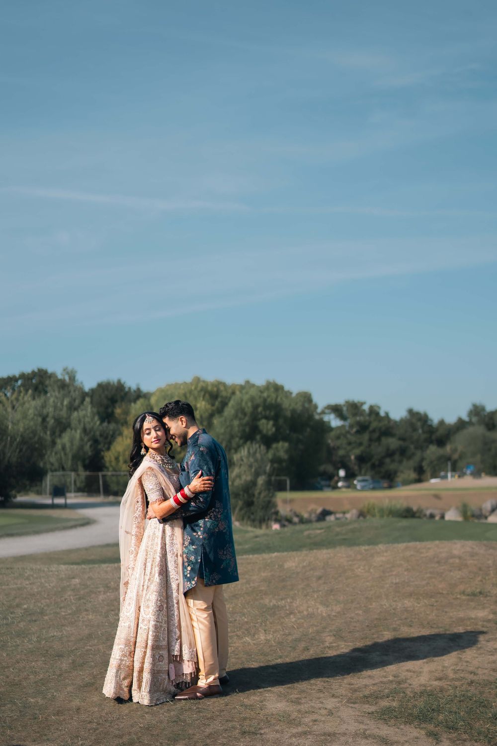 South Asian couple in traditional attire embracing outdoors, bride in embroidered lehenga and groom in teal sherwani.