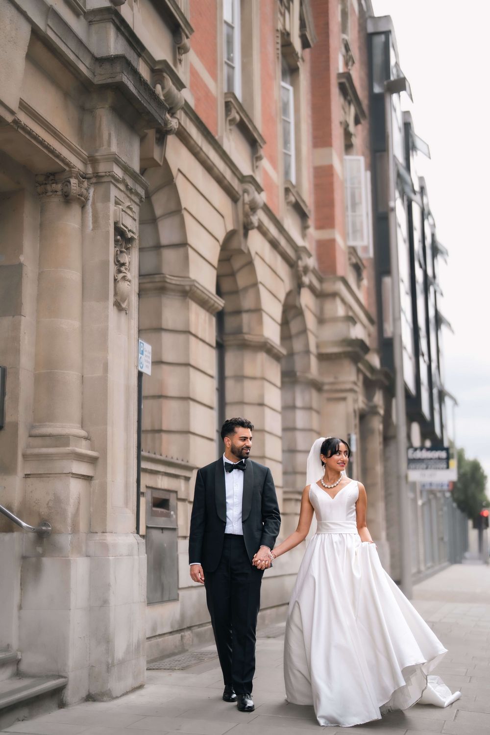 Bride and groom walk hand in hand along a classic stone building street, she in white gown, he in black tuxedo.