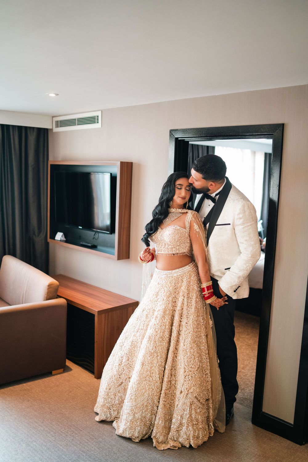 Couple in wedding attire embracing in hotel room, bride in gold lehenga and groom in white tuxedo jacket.