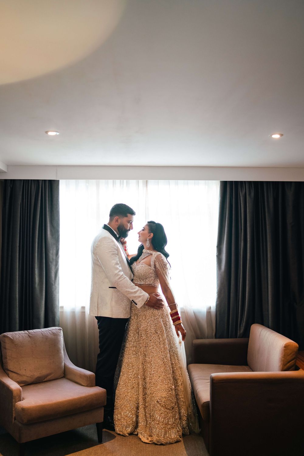 Indian bride and groom in elegant wedding attire share a romantic moment in a softly lit hotel suite.