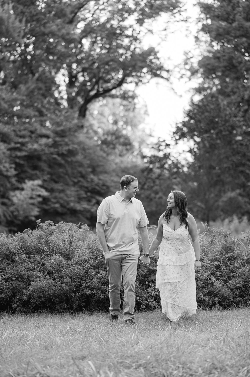 Black and white photo of a couple holding hands and walking through a lush green park, smiling at each other.