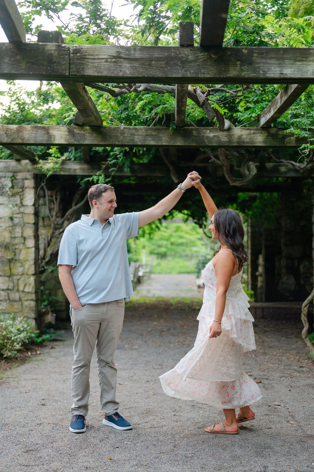 Man in blue polo twirling woman in floral dress under rustic wooden pergola in garden setting.