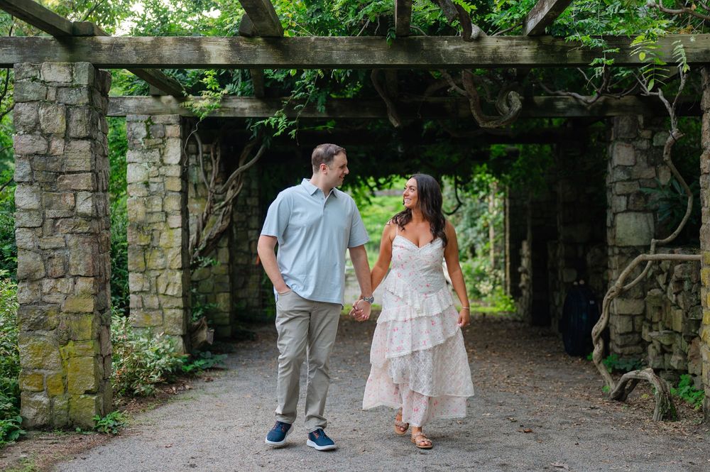 Couple holding hands and walking under a vine-covered stone pergola in a lush garden setting.