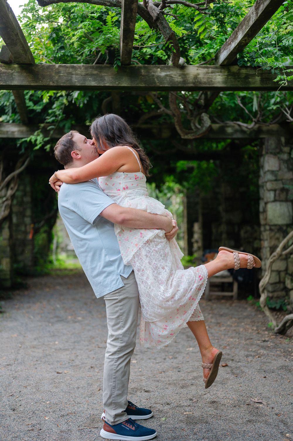 Man lifting and kissing woman in floral dress under vine-covered wooden pergola in lush garden.