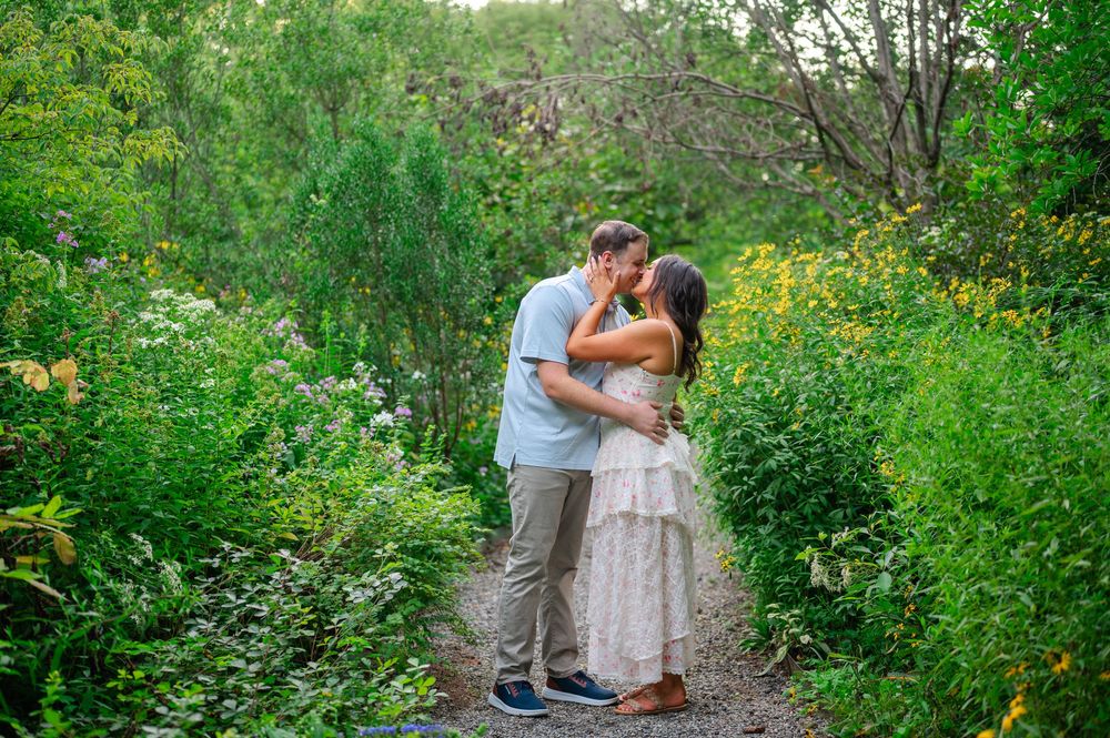 Couple sharing a romantic kiss on a garden path surrounded by lush green foliage and wildflowers.