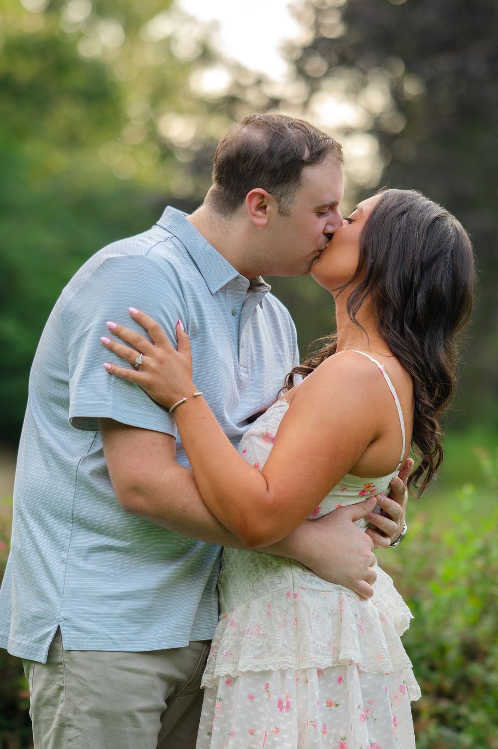 Couple sharing a romantic kiss outdoors, man in blue shirt and woman in floral dress, surrounded by lush greenery.