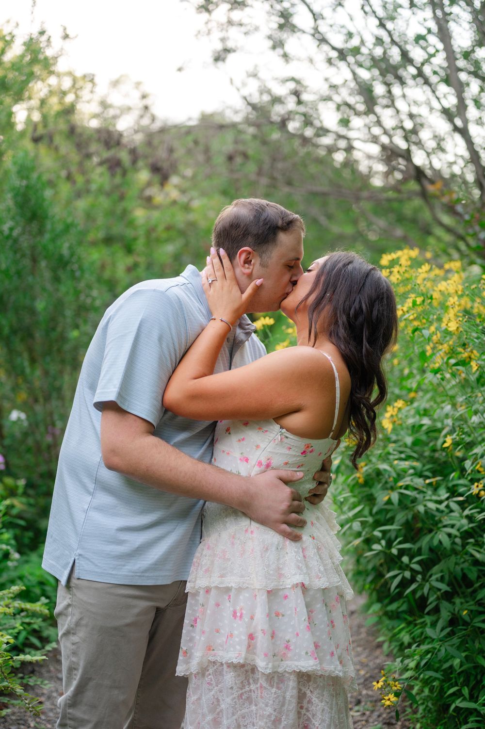 Engaged couple sharing a passionate kiss surrounded by vibrant wildflowers and greenery outdoors.